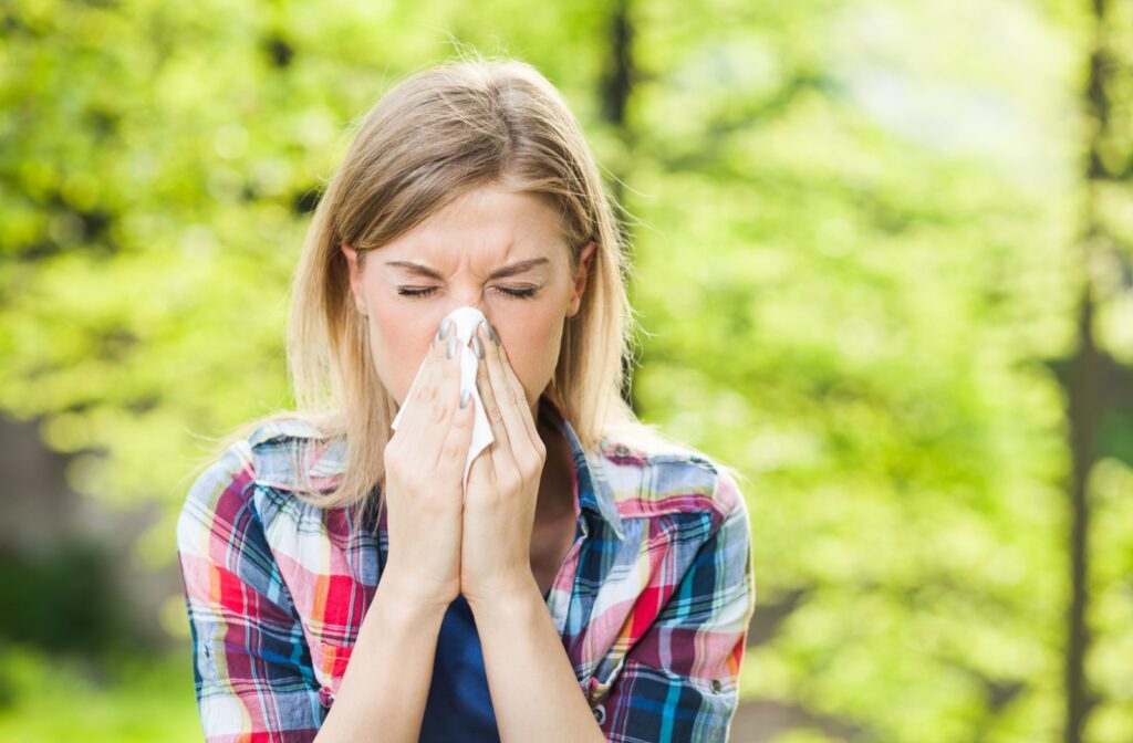 A woman is outdoors, sneezing into a tissue with her eyes closed due to her allergies.