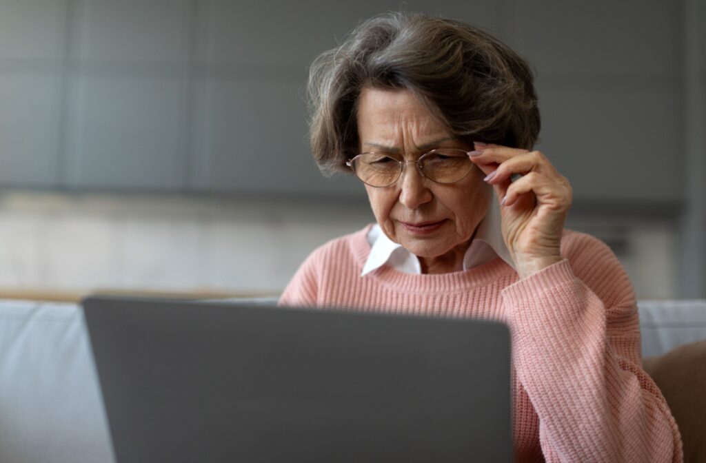 Senior woman in pink sweater squinting and holding glasses while reading off a laptop.