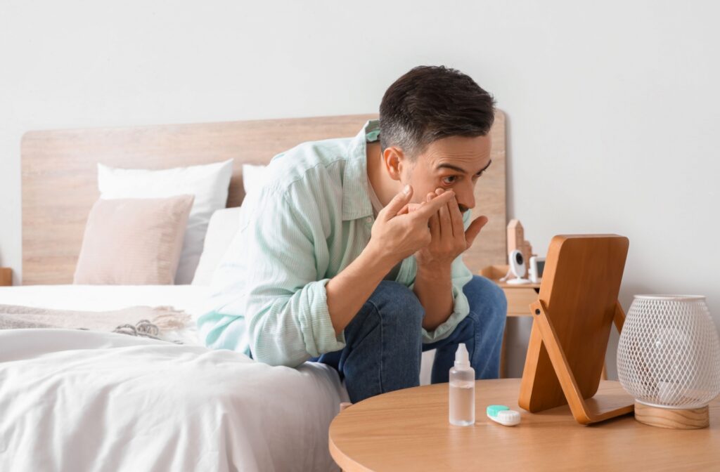 A person puts in a contact lens while sitting on a bed with a mirror and lens case nearby.