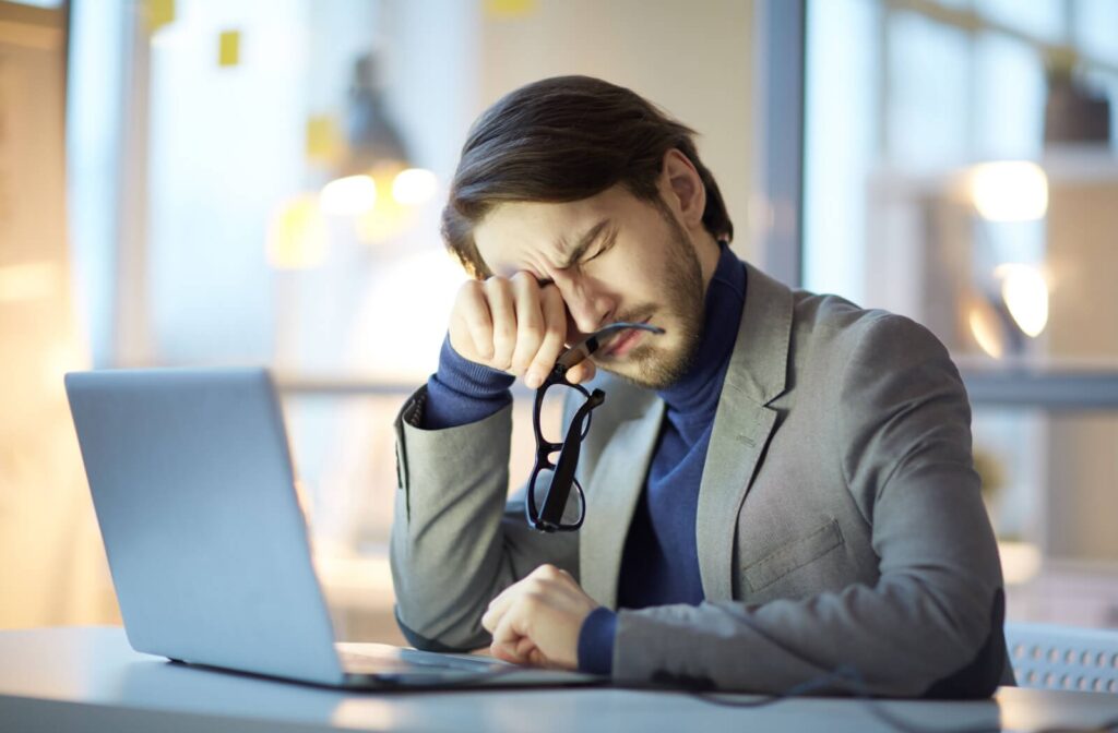 A person sitting at a desk in front of a laptop, holding a pair of glasses and rubbing their eyes due to blurry vision.