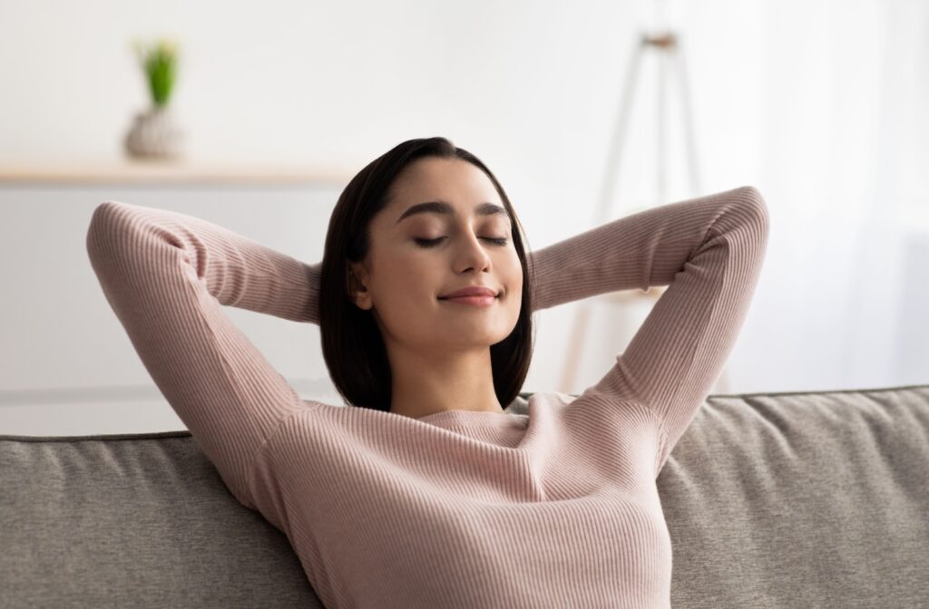 A person sitting on the couch with their eyes closed and hands behind their head, taking a moment to rest their eyes after experiencing blurry vision.