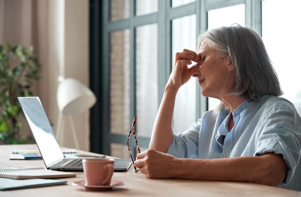 A woman working on a laptop, holding her eyes due to dry eye symptoms.