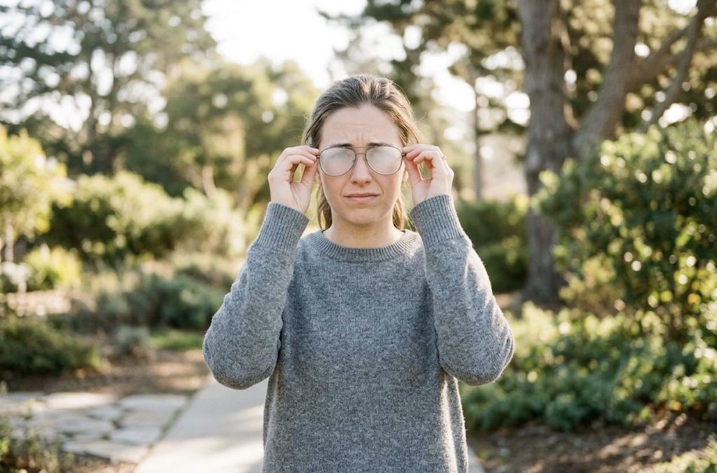 An adult stands on an outdoor path holding their foggy eyeglasses with both hands, looking frustrated.